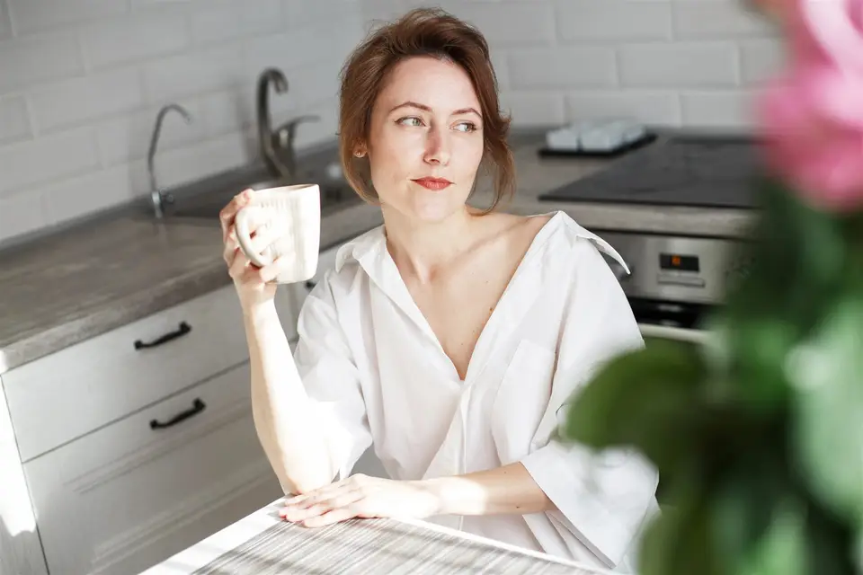 Femme pensive, une tasse à la main, assise à la table de sa cuisine