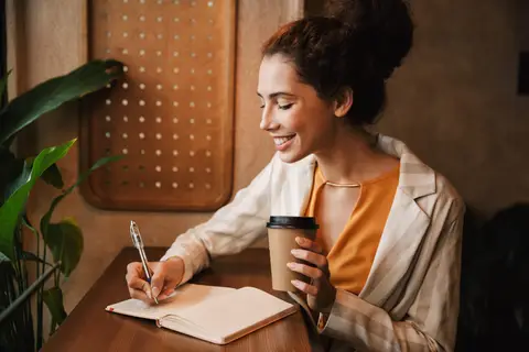 Femme souriante qui écrit dans un carnet, un café à la main