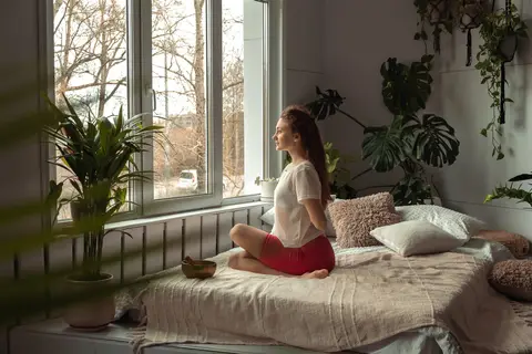 Femme assise en tailleur qui médite sur son lit, près d'une fenêtre