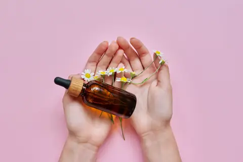 Mains qui tiennent un flacon d'huile et des fleurs de camomille sur fond rose