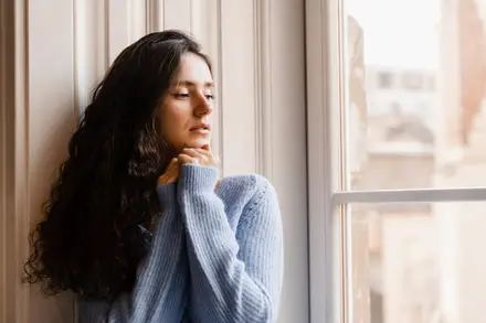 Femme pensive en pull bleu, adossée près d'une fenêtre lumineuse